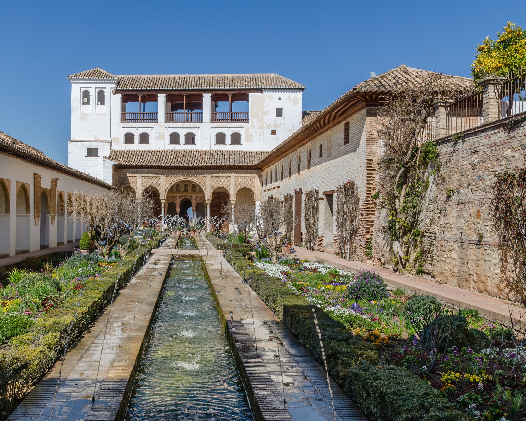 The gardens of the Generalife on the hillside above the Alhambra, with a long rectangular reflecting pool flanked by clipped cypress hedges and rose beds, with the Nasrid towers of the main palace visible in the distance.
