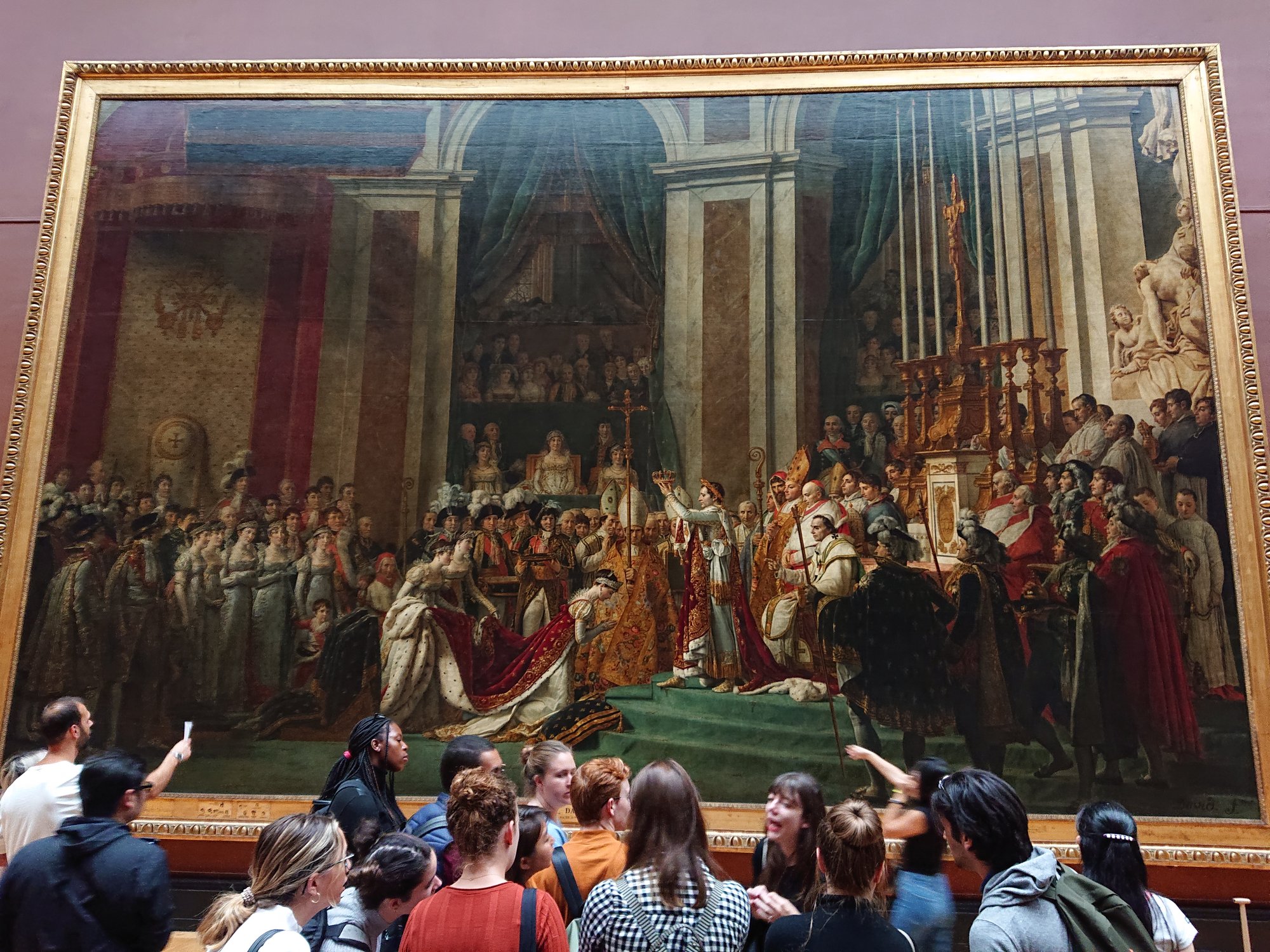 Tourists crowd in front of the giant gilt-framed coronation painting on a red gallery wall, several pointing up at the canvas.