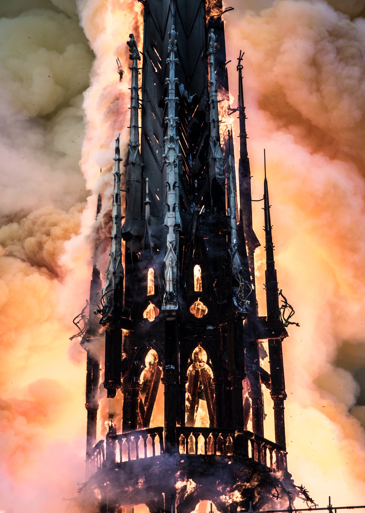 A vertical photograph of the central spire of Notre-Dame on fire on April 15, 2019, with flames consuming the wooden flèche and smoke billowing into the sky.