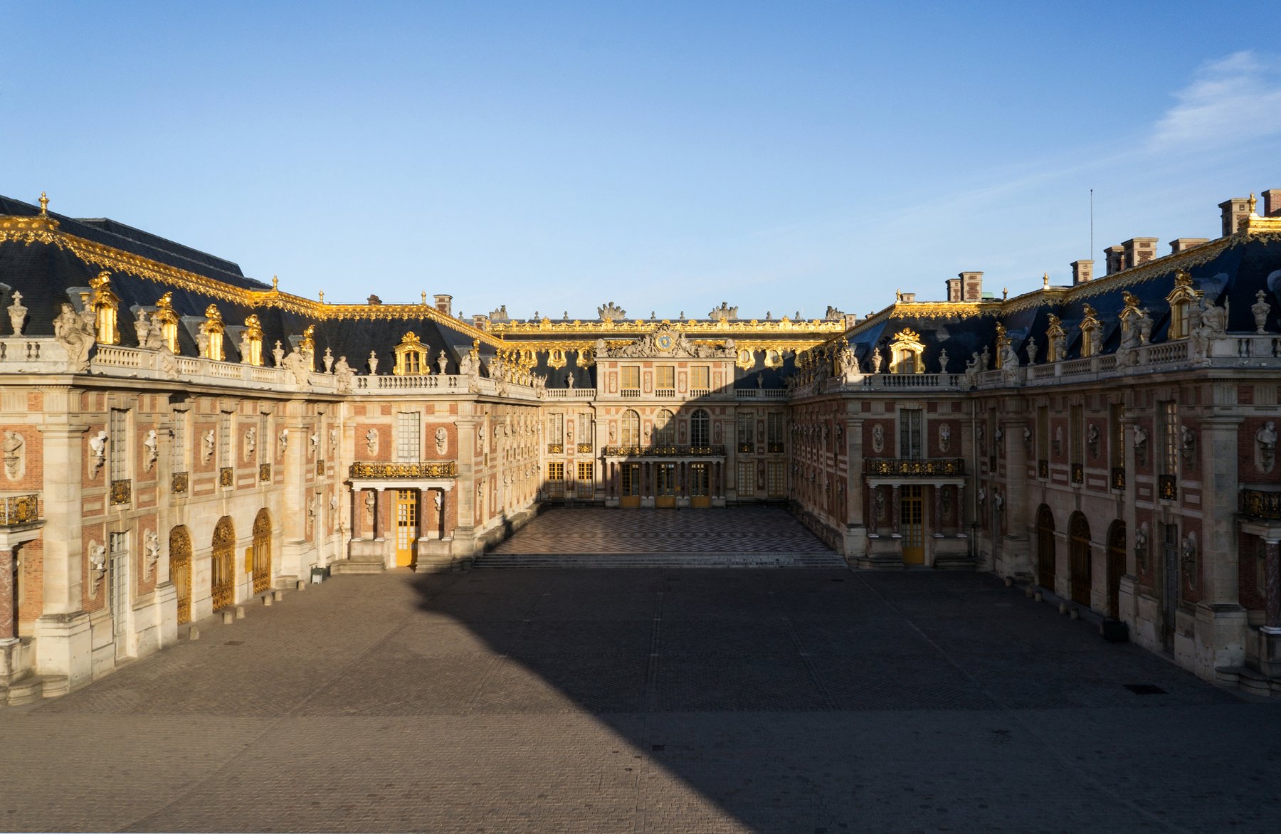 Aerial view of the Palace of Versailles, showing the long horseshoe facade of the central palace, the formal gardens stretching west toward the Grand Canal, and the surrounding parkland of the royal domain.