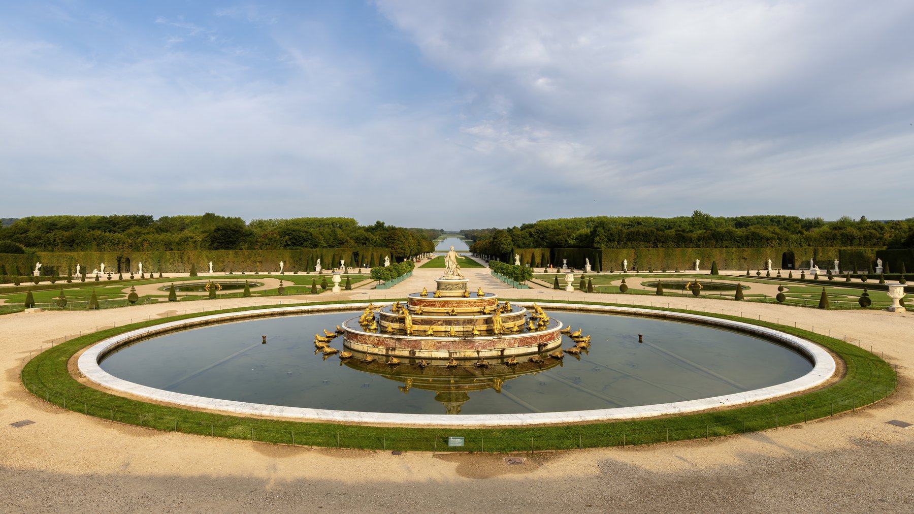A panoramic view of the gardens of Versailles seen from the western terrace of the palace, with the Latona Fountain in the center, parterres of clipped boxwood and grass, and the Grand Canal stretching toward the horizon.