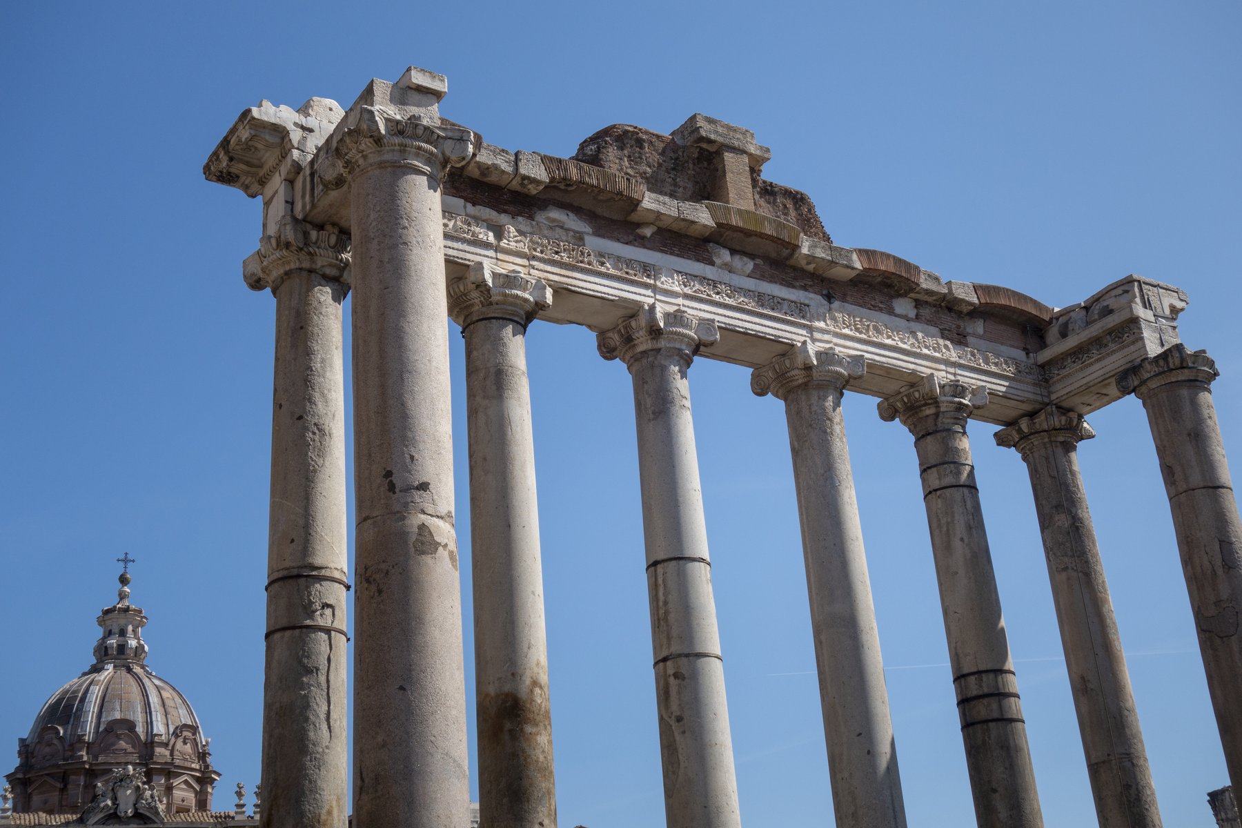 The eight surviving columns of the Temple of Saturn in the Roman Forum, standing in a row against a clear blue sky with the brick remains of other forum structures around them.