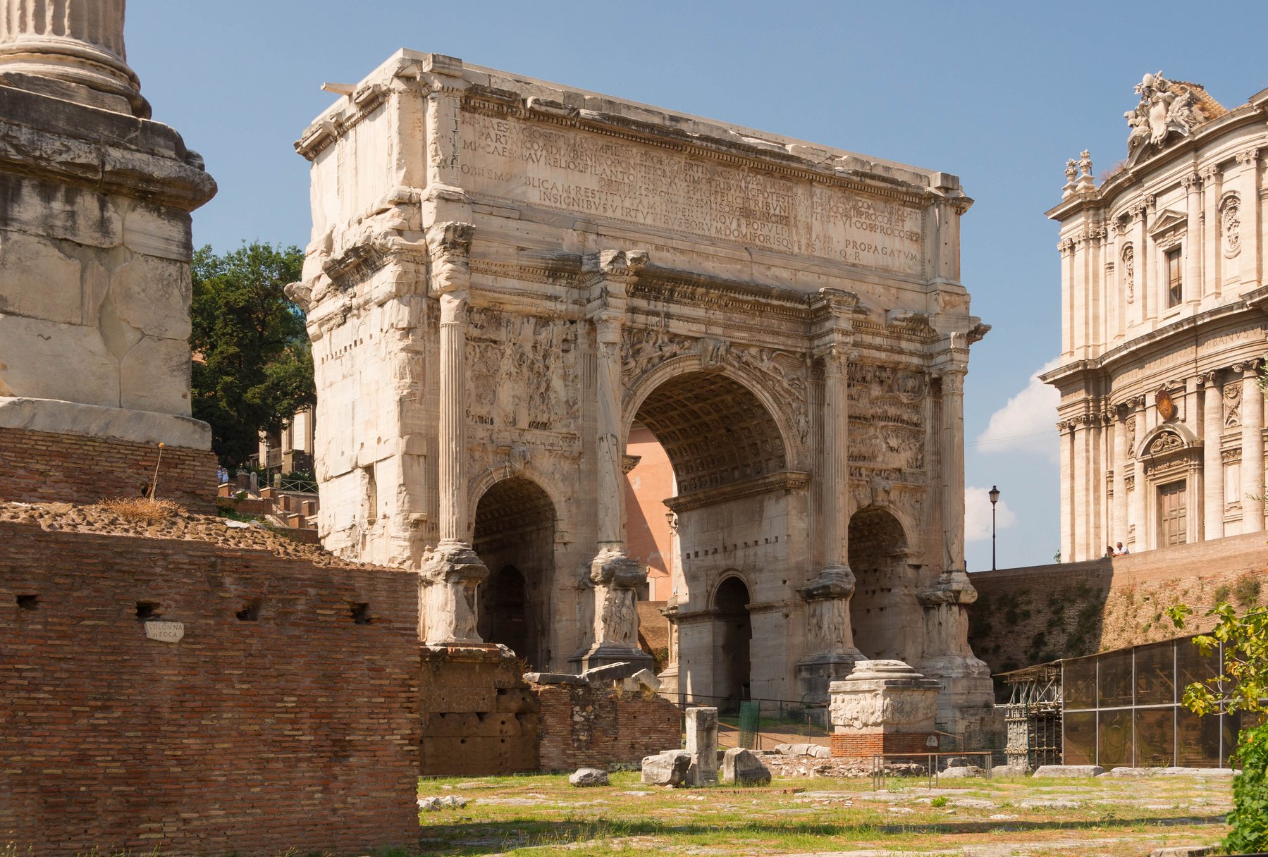 The Arch of Septimius Severus at the western end of the Roman Forum, a triple-bay triumphal arch of white marble with carved reliefs showing the emperor's military campaigns, the Column of Phocas standing nearby.