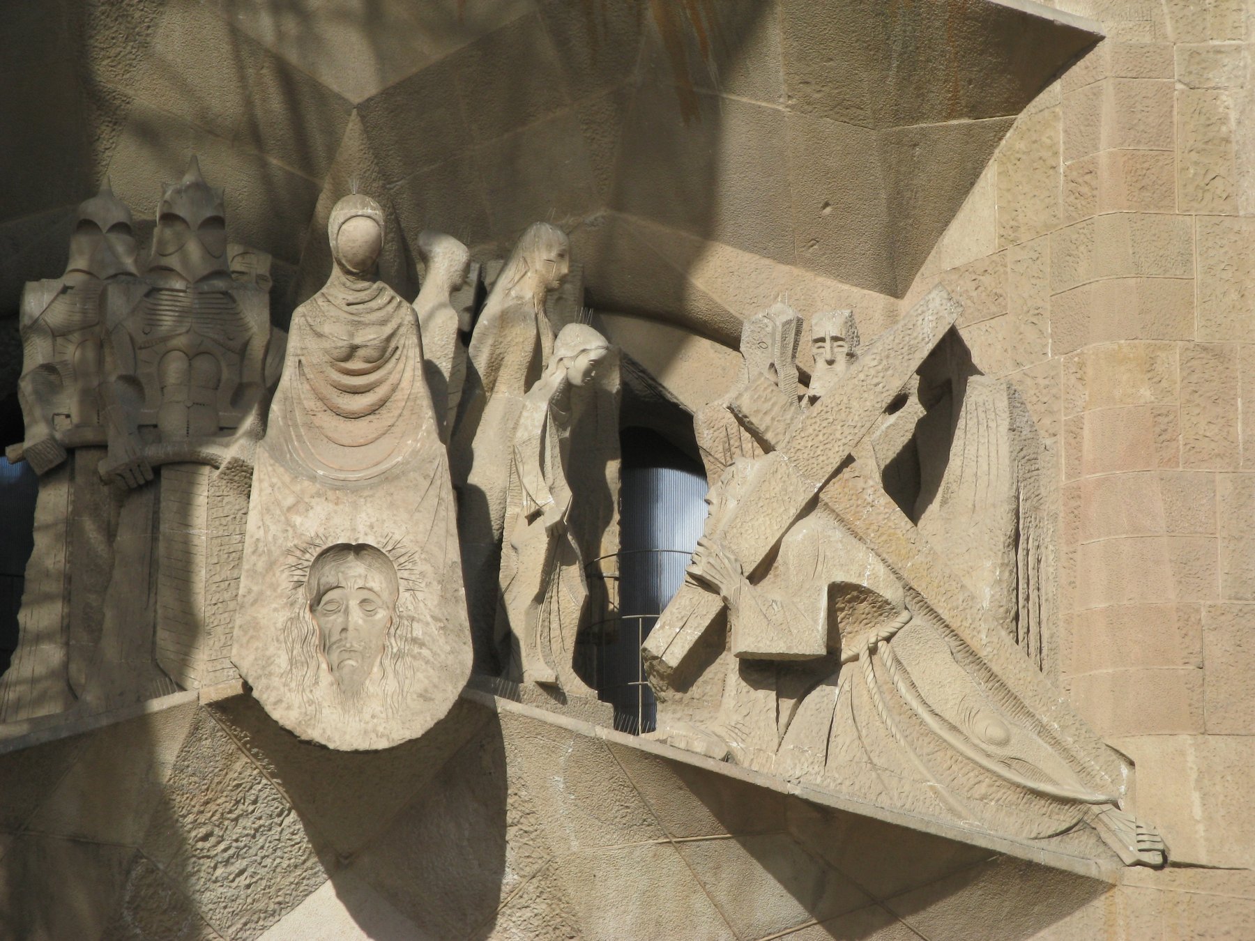 The Passion facade of the Sagrada Familia by Josep Maria Subirachs, with severe angular stone figures depicting the suffering of Christ on a porch of slanted columns, in a starkly different style from the Nativity facade.