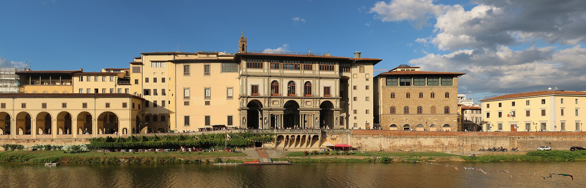 The Uffizi Gallery seen from the Arno river, the long facade of the building running up from the river toward the Palazzo Vecchio's tower, with the Vasari Corridor visible as a covered arched passage running across the bottom of the wall along the Lungarno.